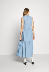 Lichtblauwe denim maxi-jurk met een gelaagde rok, mouwloos ontwerp en een gesloten halslijn. Voorzien van een subtiele ruchedetail bij de kraag.