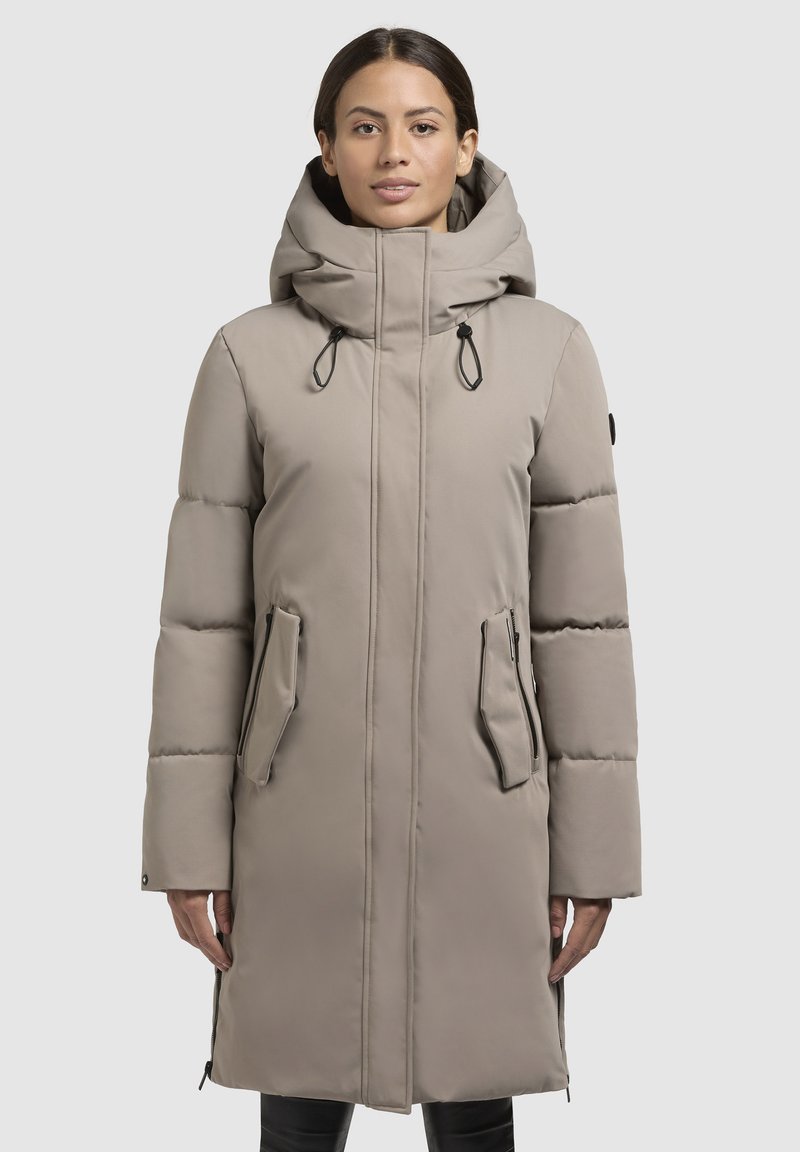 Langer beiger Puffer-Mantel mit Kapuze, ausgestattet mit einem Frontreißverschluss, verstellbaren Kordelzügen und zwei seitlichen Taschen mit Klappen. Glatte Textur.