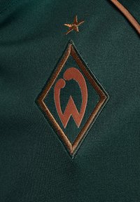Broderad logotyp på en mörkgrön tyg med en brun diamantform med stiliserade bokstäver och en liten stjärna ovanför.