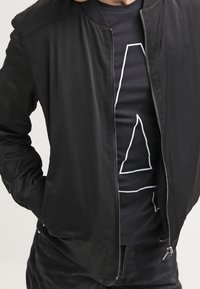 Musta bomber-takki, jossa on sileä kangas, resorikaulus ja vetoketjullinen etuosuus. Pukeutunut mustan t-paidan päälle, jossa on valkoinen viivakuosi.