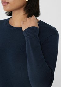 Marineblauer Strickpullover mit Rundhalsausschnitt, gerippten Bündchen und strukturiertem Stoff. Nahaufnahme einer Hand, die auf der Schulter ruht.