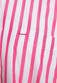 Camisa de rayas con llamativas rayas rosas y blancas, de material de algodón, con una pequeña costura de acento roja en el bolsillo. Textura suave.