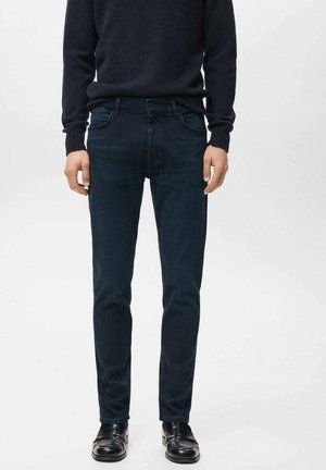 Jeans azul oscuro con un diseño de ajuste ceñido, confeccionados en denim con una textura sutil. Combinados con un suéter oscuro y zapatos negros.