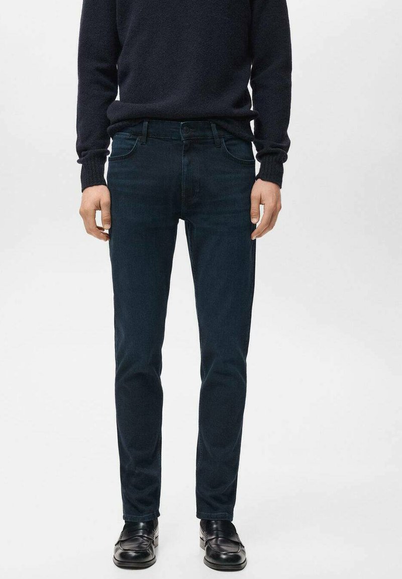 Jeans azul oscuro con un diseño de ajuste ceñido, confeccionados en denim con una textura sutil. Combinados con un suéter oscuro y zapatos negros.