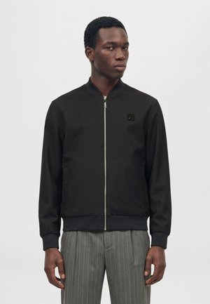 Un homme pose avec une veste noire zippée ornée d’un logo sur la poitrine et un pantalon gris à fines rayures, sur un fond blanc uni.