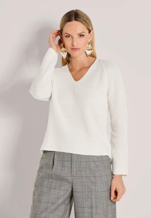 Vrouw met steil blond haar draagt een witte gebreide trui, grote geometrische gouden oorbellen en grijze geruite broek, staand tegen een neutrale muur.