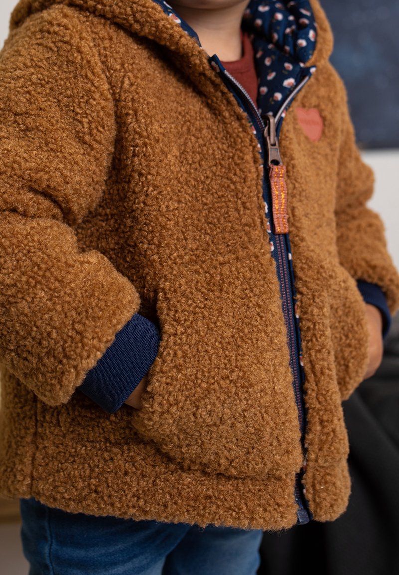 Enfant portant une veste marron duveteuse avec les mains dans les poches sur un sweat à capuche bleu marine à motif floral et un jean en denim.