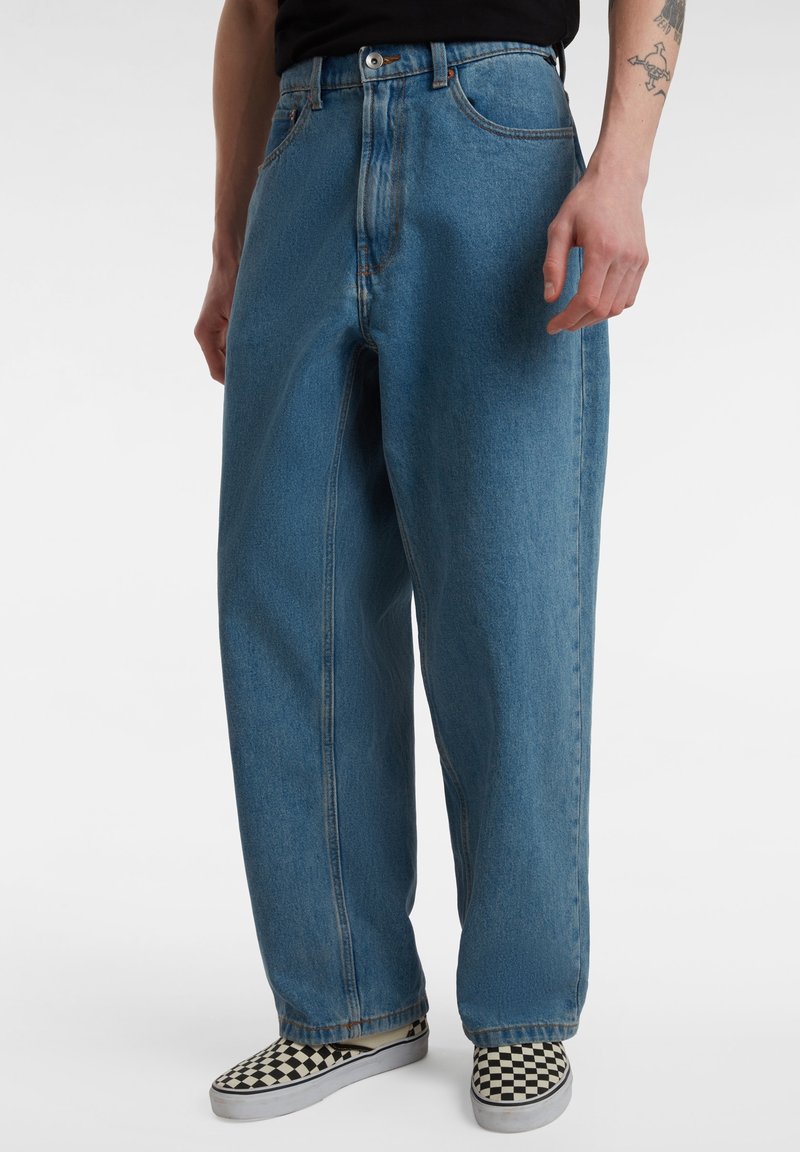 Personne portant un jean bleu ample, des chaussures à enfiler à carreaux noirs et blancs, et un t-shirt noir, avec un tatouage visible sur l'avant-bras droit.