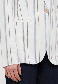 White linen blazer with light blue and brown vertical stripes. Features a single button closure and side pocket. Worn over dark denim.