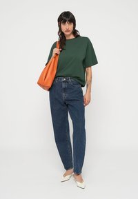 Πράσινο υπερμεγέθες t-shirt, μπλε τζιν με υψηλή μέση, λευκά μυτερά mules και πορτοκαλί τσάντα ώμου, όλα πάνω σε λευκό φόντο.