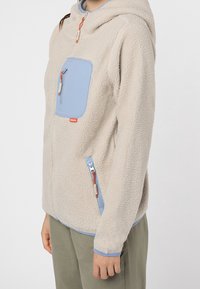 Fleece hoodie in lichtbeige, met een blauw ritsvak op de borst, zijzakken en oranje ritsen. Zachte textuur, casual ontwerp.