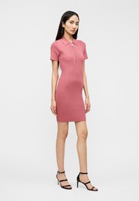 Robe rose moulante côtelée avec un col polo et une fermeture éclair à l'avant, portée avec des talons hauts noirs à brides.