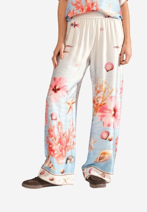 Pantalon blanc ample avec un motif sur le thème de l'océan, comprenant des coraux, des coquillages, des étoiles de mer, des fleurs roses et de petits poissons, assorti à des chaussures foncées.