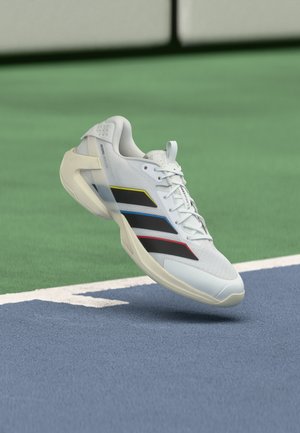 Witte sportschoen met zwarte strepen en gekleurde randen zwevend boven een blauwgroen tennisveld.