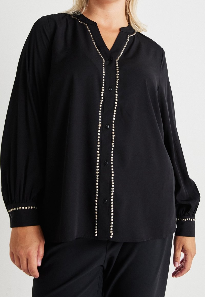 Blouse noire à manches longues boutonnée avec des détails en clous argentés le long de la boutonnière et des poignets portée par une personne à la peau claire et aux cheveux blonds.