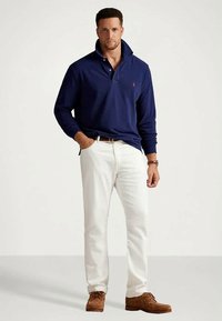 Mężczyzna w granatowej koszulce polo z długim rękawem, białych spodniach, z brązowym paskiem i butami, stoi z jedną ręką w kieszeni na jednolitym tle.