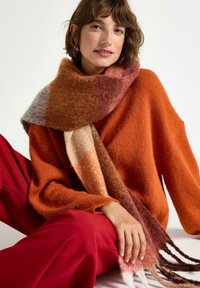 Gehaakte sjaal met gemengde warme tinten van oranje, roze en bruin; zachte textuur met franjes aan de uiteinden. Draag het met een gezellige oranje trui en rode broek.