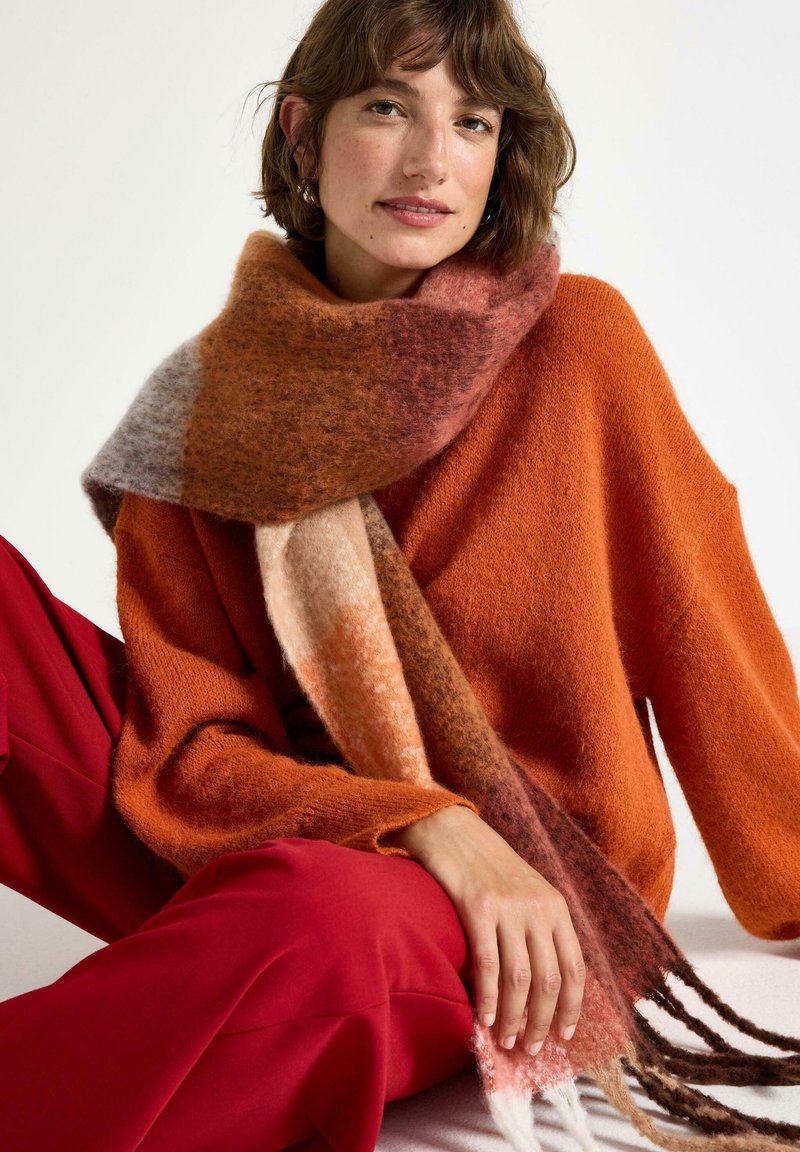 Gehaakte sjaal met gemengde warme tinten van oranje, roze en bruin; zachte textuur met franjes aan de uiteinden. Draag het met een gezellige oranje trui en rode broek.