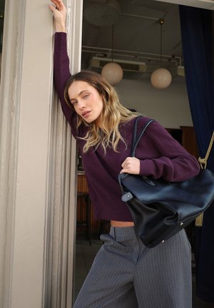 Paarse gebreide trui, gestreepte grijze broek en een zwarte leren schoudertas. De tas heeft een minimalistisch ontwerp met een omvouwflap en zilverkleurige hardware.