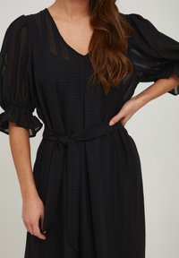 Černé šaty se strukturou, V-výstřihem, nařasenými rukávy a uvázaným páskem v pase. Mají vrstvený, splývavý design s jemným vzorem.