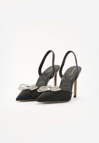 Escarpins à talon aiguille en tissu noir avec bouts pointus, brides arrière et un accent en nœud orné de bijoux. Texture lisse avec un léger brillant sur le dessus.