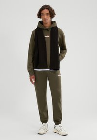Zestaw oliwkowej zielonej bluzy z kapturem i spodni z czarnym dzianinowym szalikiem. Biały logo na bluzie. Białe trampki z czarnymi akcentami. Casualowy, luźny krój.