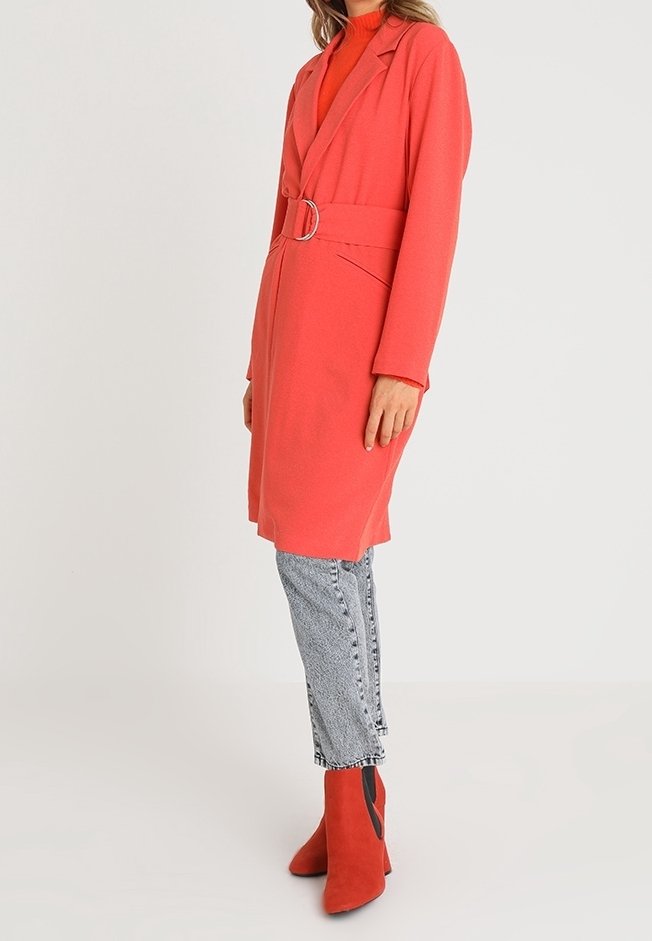 Femme portant un manteau corail ceinturé sur un jean gris, associé à des bottines corail, debout devant un fond clair uni.