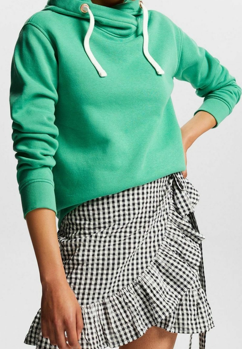 Femme portant un sweat à capuche vert avec des cordons blancs et une jupe à volants à carreaux noir et blanc, montrée du cou jusqu'à mi-cuisse.