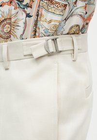 Κοντινό πλάνο σε κρεμ παντελόνι με υφασμάτινη ζώνη και ασημένια αγκράφα που φοράται με κορυφή με μοτίβα, με σχέδια ήλιου και λουλουδιών.