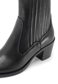 Botín de cuero negro hasta el tobillo con detalles acanalados, tacón bajo y textura suave; presenta cortes curvados y costuras en contraste.