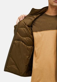 Giacca a vento marrone con texture trapuntata, aperta per rivelare una maglia a strati marrone e beige sottostante. Tessuto liscio con un sottile contrasto di colore.