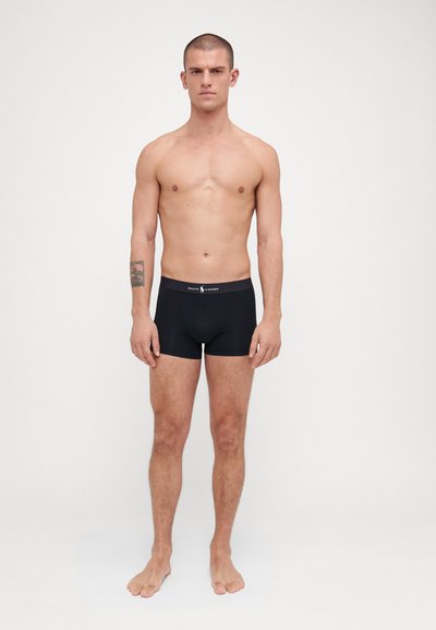Boxer shorts noirs en tissu extensible avec une texture lisse. Dotés d'une ceinture avec logo et d'un design ajusté. Le modèle est debout pieds nus.