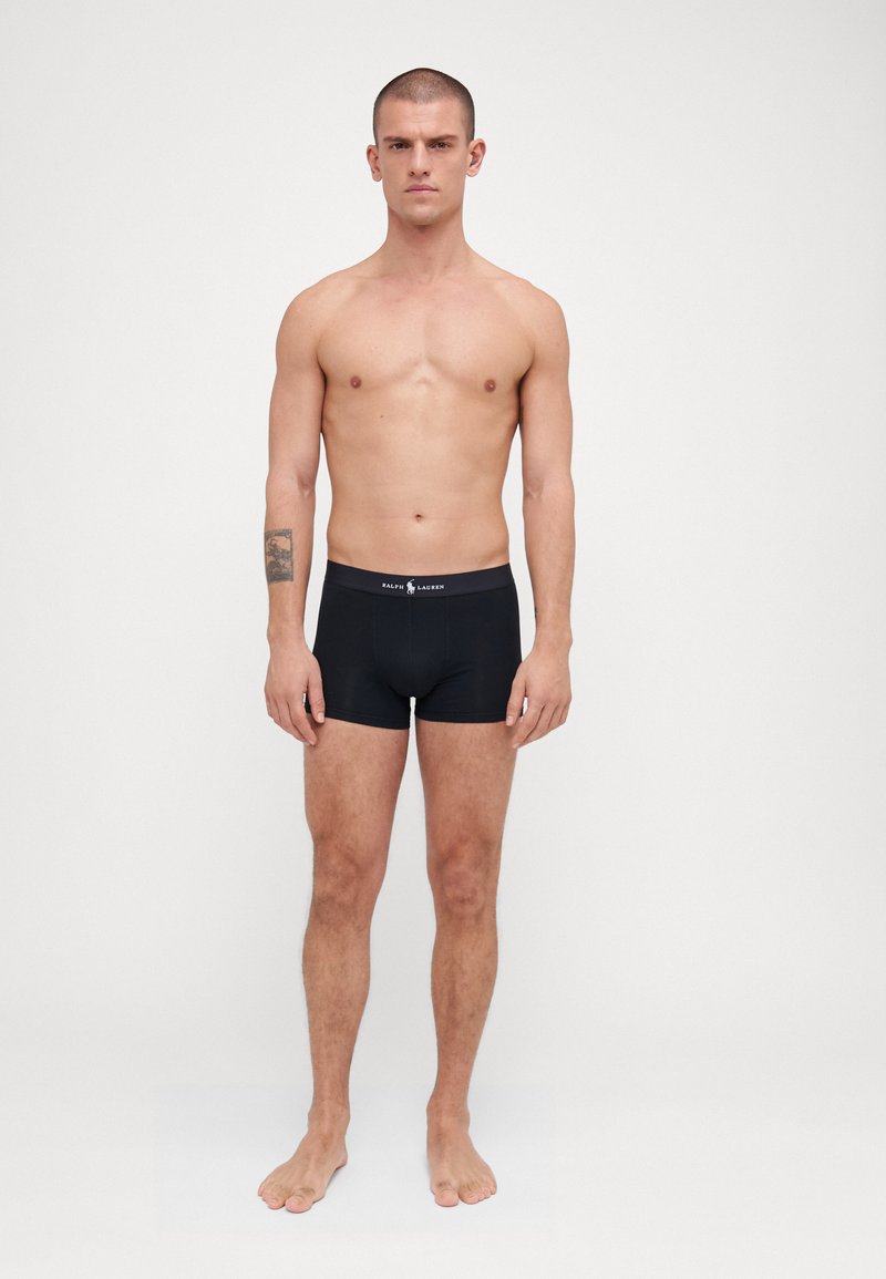 Boxer shorts noirs en tissu extensible avec une texture lisse. Dotés d'une ceinture avec logo et d'un design ajusté. Le modèle est debout pieds nus.