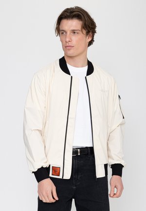 Mann trägt eine cremefarbene Bomberjacke mit schwarzem Kragen und Bündchen, weißem T-Shirt, schwarzer Jeans und einem schwarzen Gürtel vor einem einfarbigen Hintergrund.