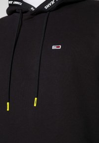 Czarna bluza z kapturem, z małym logo w czerwonych, białych i niebieskich akcentach. Regulowane sznurki z żółtymi końcówkami. Miękka tekstura.