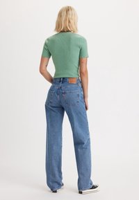 Top verde de manga corta con textura acanalada, combinado con unos pantalones vaqueros de pierna ancha de talle alto en un tono azul claro. Parche de cuero notable en la parte trasera de la cinturilla. Zapatillas negras visibles.