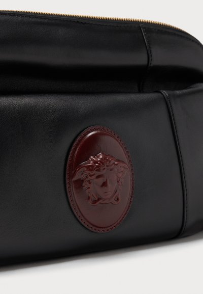 Μαύρη δερμάτινη τσάντα με καφέ οβάλ έμβλημα Μέδουσας ανάγλυφο στο μπροστινό μέρος και ορατό χρυσό φερμουάρ κατά μήκος του πάνω άκρου.