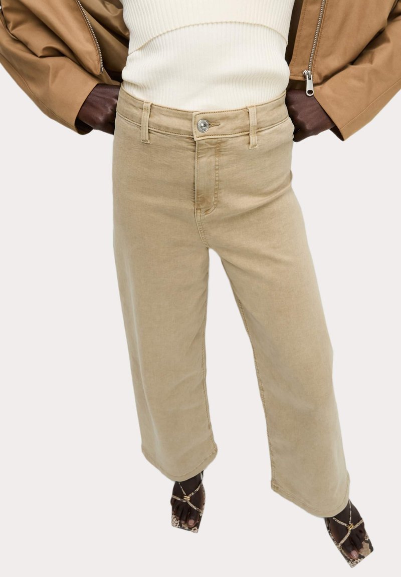 Béžové široké džíny z měkkého denimu s klasickými předními kapsami a kovovým knoflíkem. Spárovány s krémovým ribbed topem a hnědou koženou bundou.