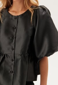Top in satin nero con scollo rotondo, maniche a sbuffo e chiusura frontale con bottoni. Presenta una vita arricciata e una texture liscia.