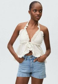 Top bianco con scollo a halter, bordi arricciati e legatura frontale, abbinato a pantaloni shorts in denim blu chiaro. Il tessuto sembra leggero e texturizzato.