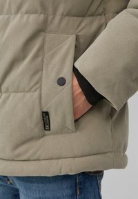 Die Hand einer Person in der Tasche einer beige gesteppten Jacke mit schwarzem Druckknopf und Stoffetikett, kombiniert mit blauen Jeans.