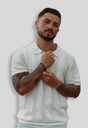 White short-sleeve polo shirt with textured zigzag pattern; model poses with hand on sleeve, showcasing detailed tattoos and a black bracelet.