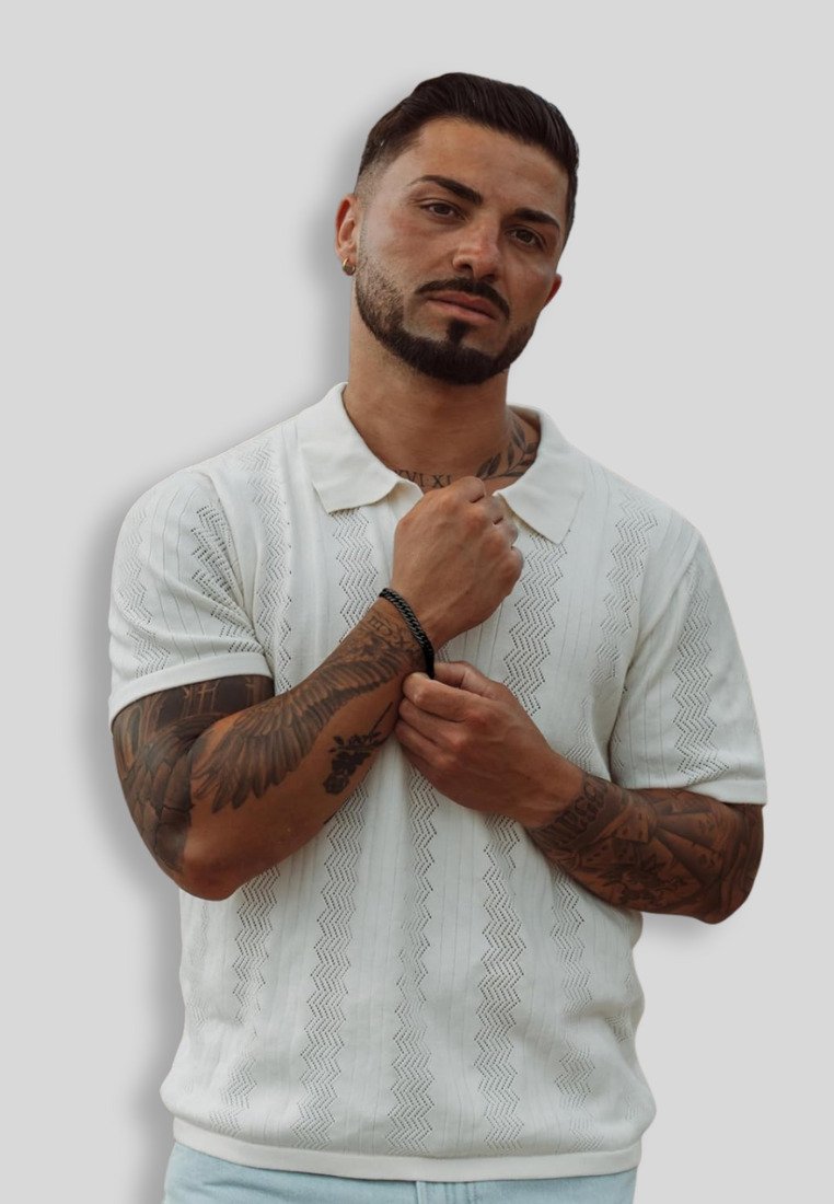 White short-sleeve polo shirt with textured zigzag pattern; model poses with hand on sleeve, showcasing detailed tattoos and a black bracelet.