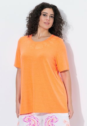 Kvinde med krøllet hår iført en orange kortærmet top med subtile skaldyrsdesign nær udskæringen og hvide bukser med lyserøde mønstre.