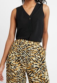 Černý bezrukávový top s V- výstřihem a detaily knoflíků, kombinovaný s kalhotami s tigerovým vzorem v žluté a černé barvě, které mají elastický pas a boční kapsy.