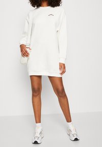 Vestido sudadera blanco con mangas largas, escote redondo y pequeño logo bordado. Combinado con zapatillas y un pequeño bolso cruzado.
