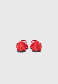 Ballerines en maille rouge avec une finition brillante, la vue arrière montre un design ouvert, une bride de cheville et des semelles noires. Texture lisse avec un motif en treillis détaillé.