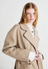 Abrigo de lana beige con cuello muesca, corte oversize, bolsillos laterales y cierre de botones, superpuesto a una camisa blanca con puños con volantes.