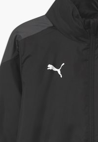 Musta kevyt takki korkean kauluksen kanssa, jossa on vetoketjusuljenta, teksturoitu kangas ja valkoinen Puma-logo vasemmassa rinta-alueessa.