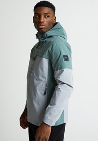 Veekindel jope rohelises ja hallis, millel on lukuga esi, reguleeritav kapuuts ja logo plekk varrukal. Sile tekstuur.